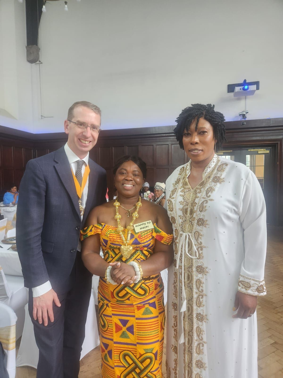 Three people pose indoors. A man in a suit stands left of two women in patterned dresses. They are smiling in a well-lit room.