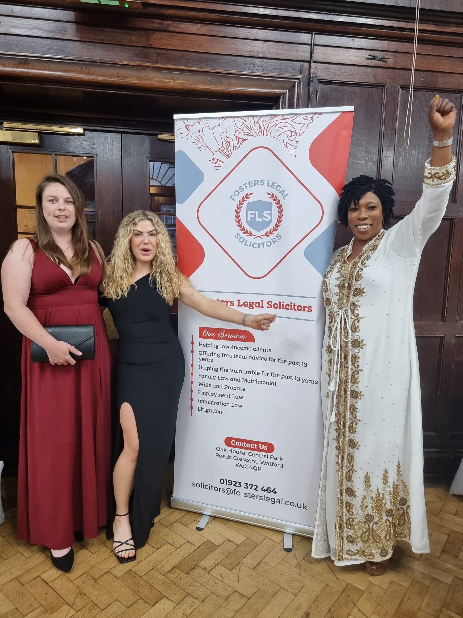 Three women pose at an event. They stand near a banner with a logo. One woman raises her fist, another points, and a third holds a clutch.