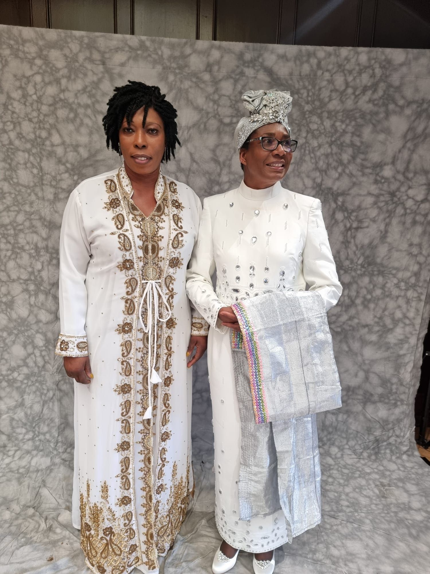Two women in white robes with head coverings pose in front of a patterned backdrop. The woman on the left has dark dreadlocks, the other wears a decorated head wrap and holds a fringed scarf.