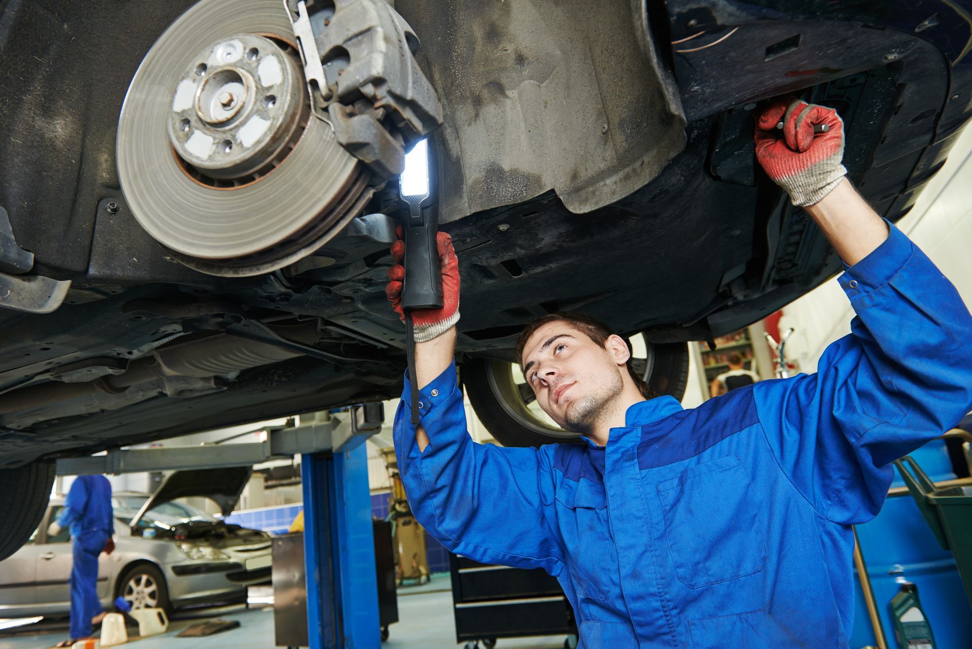 Mechanic in blue jumpsuit examining car brakes with a flashlight in a garage.