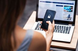 A woman is holding a cell phone in front of a laptop computer.