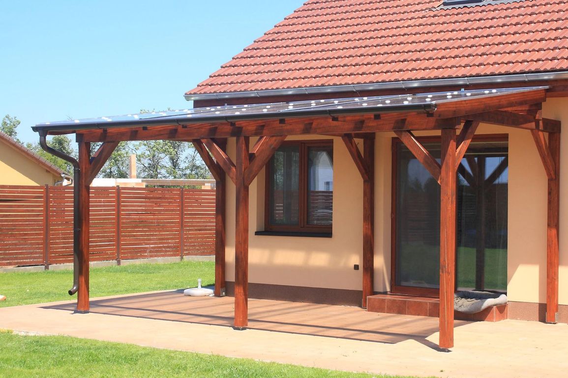 Patio coperto in legno annesso a una casa beige con tetto di tegole rosse e cortile recintato.