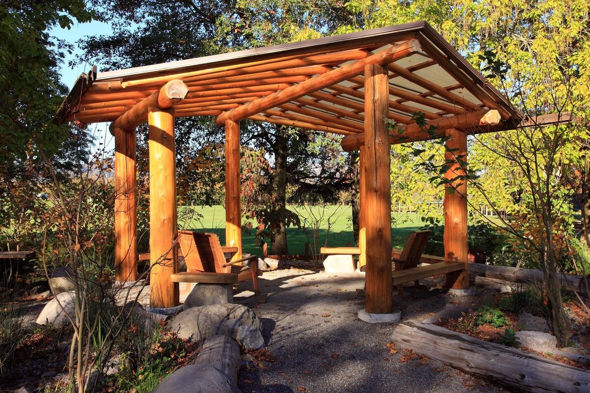 Riparo in legno per picnic con pali e panchine in un parco soleggiato circondato da rocce e alberi.