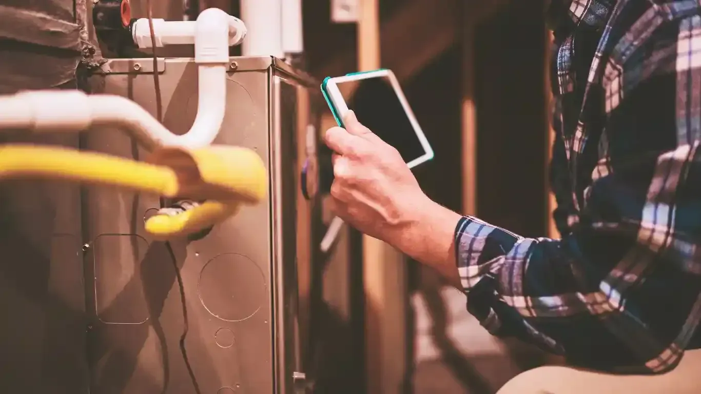 Plumber in blue shirt using multimeter to inspect heating system components in a white cabinet.