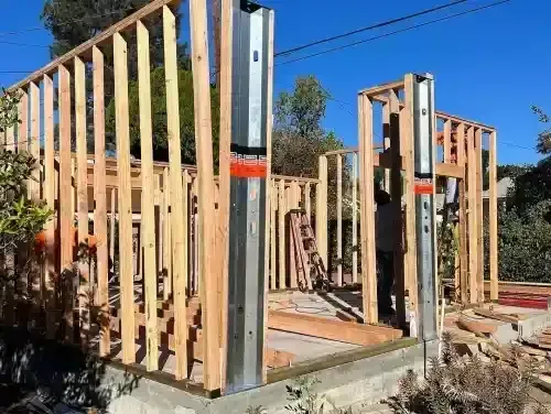 Wooden frame construction of a small building, with metal support beams and a concrete foundation, outdoors on a sunny day.