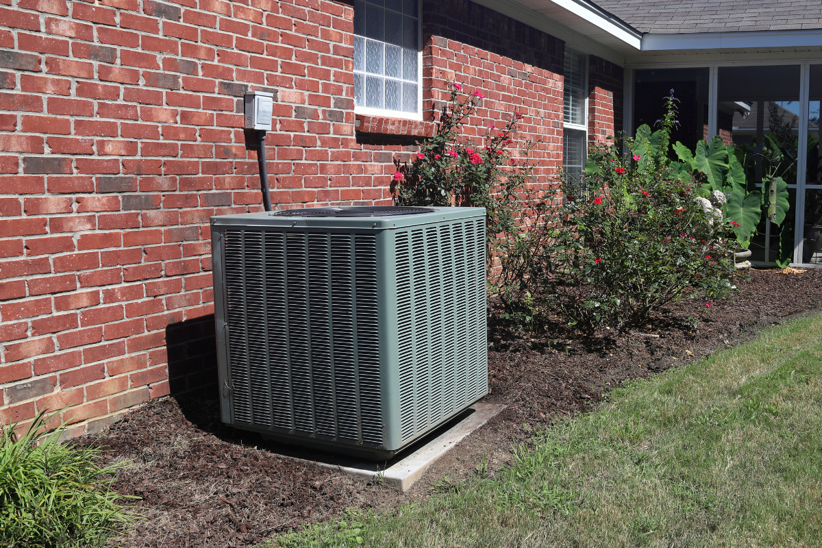 An air conditioning unit next to a brick building, set in a yard with landscaping.