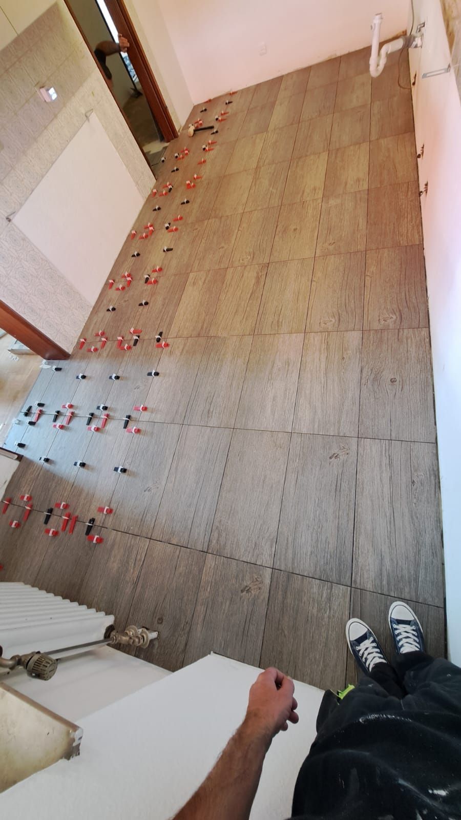Tiled wall in progress, with red tile leveling spacers. Man's legs and sneakers visible.