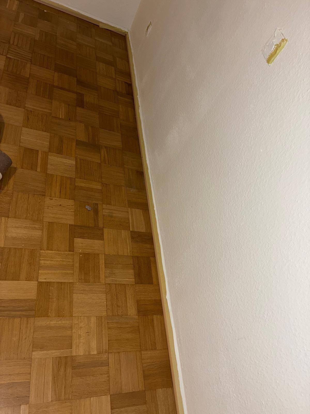 Wooden parquet flooring next to a white wall with a beige trim.