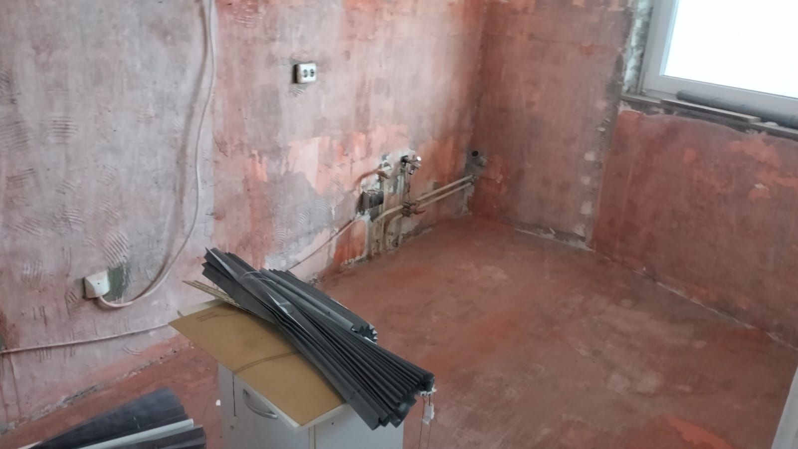 Interior view of a room under construction with exposed pipes and wiring. Reddish walls and floor, a window, and materials on the floor.