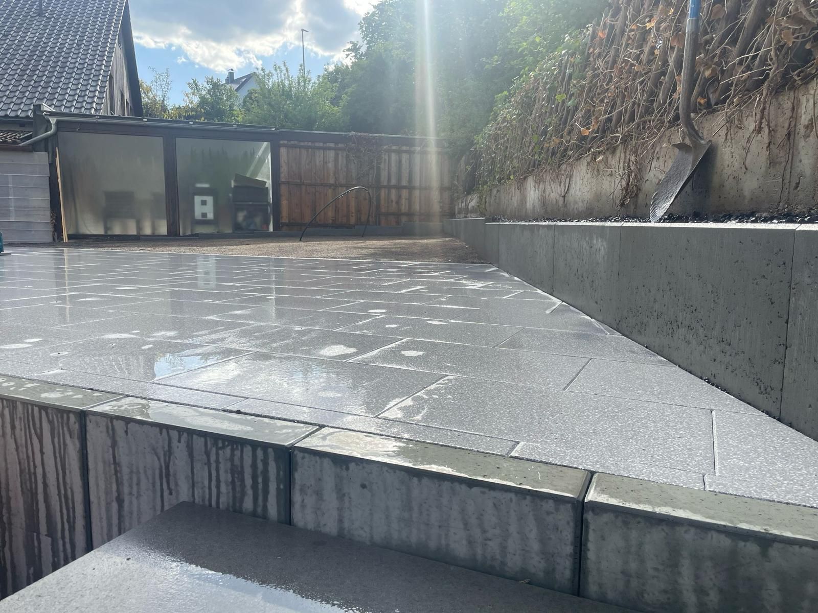 Wet concrete patio with retaining wall, sunlight, and a garage in the background.
