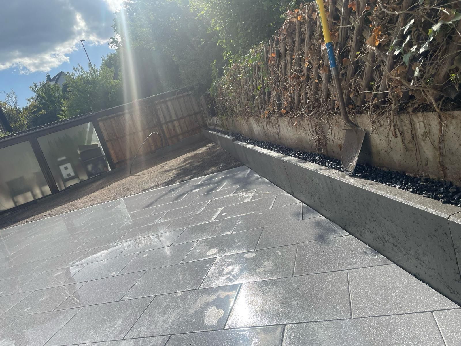 Gray tiled patio with a border of black pebbles next to a wall with shrubbery. A shovel stands nearby.