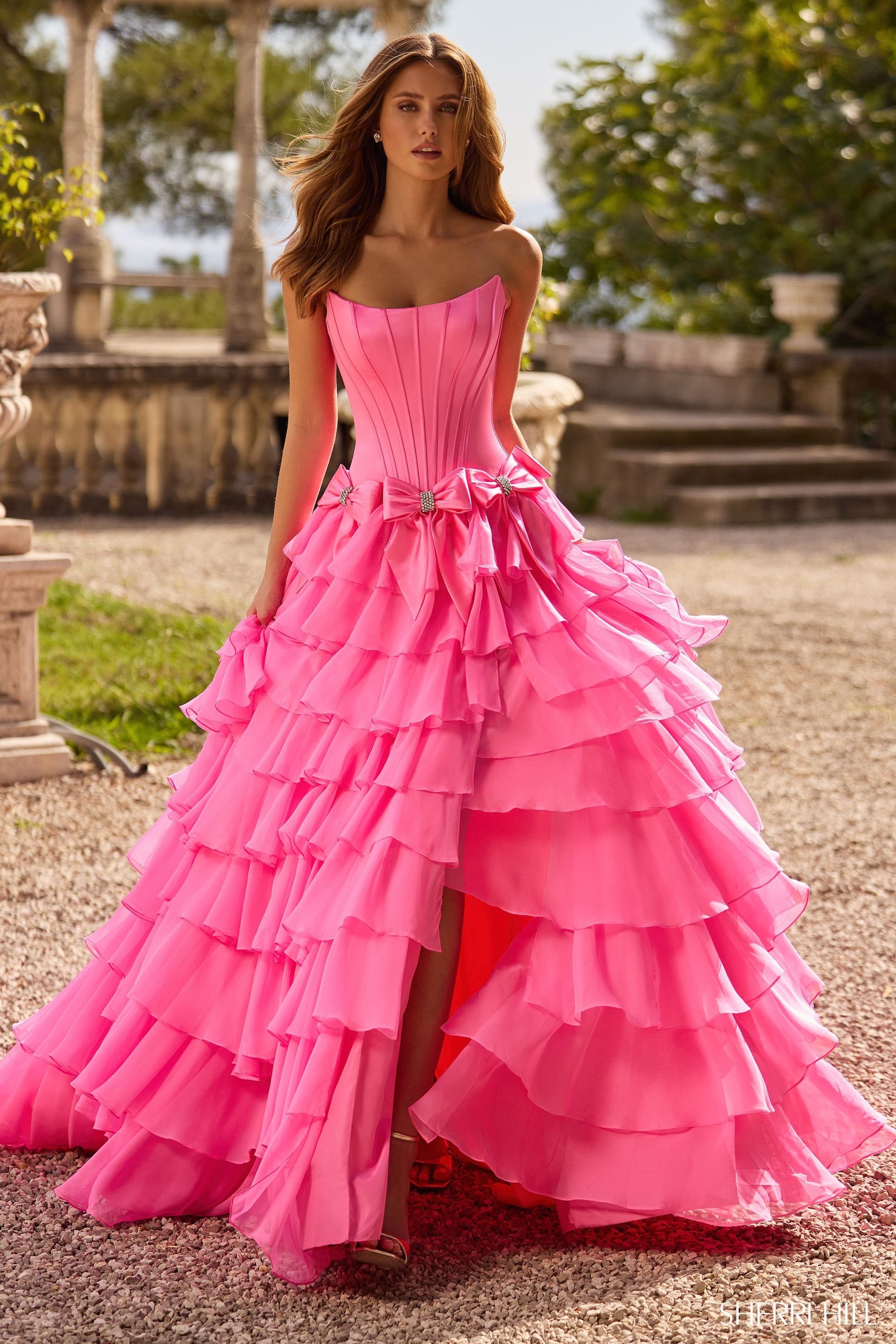 Woman in bright pink, strapless, tiered gown with high slit, standing outdoors.