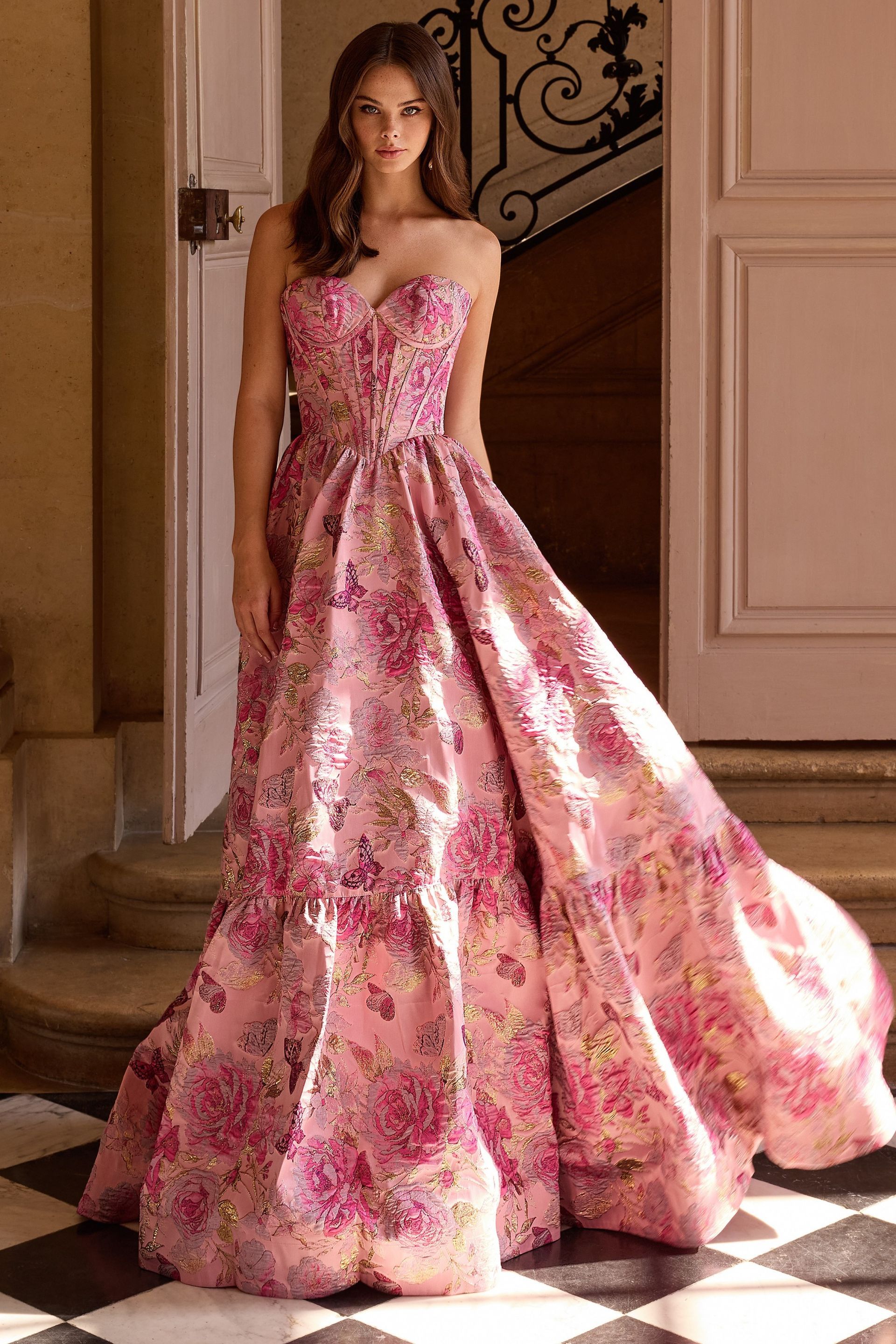 Woman in a pink floral strapless gown stands near a doorway with decorative stonework and steps.