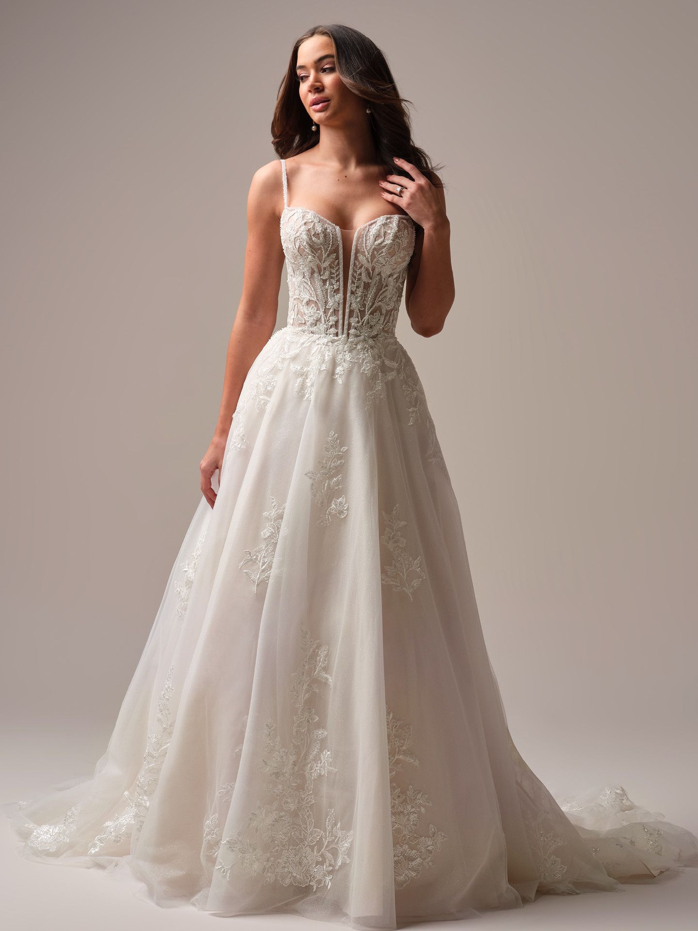 Woman in a white wedding gown with spaghetti straps, standing in a studio with a light background.