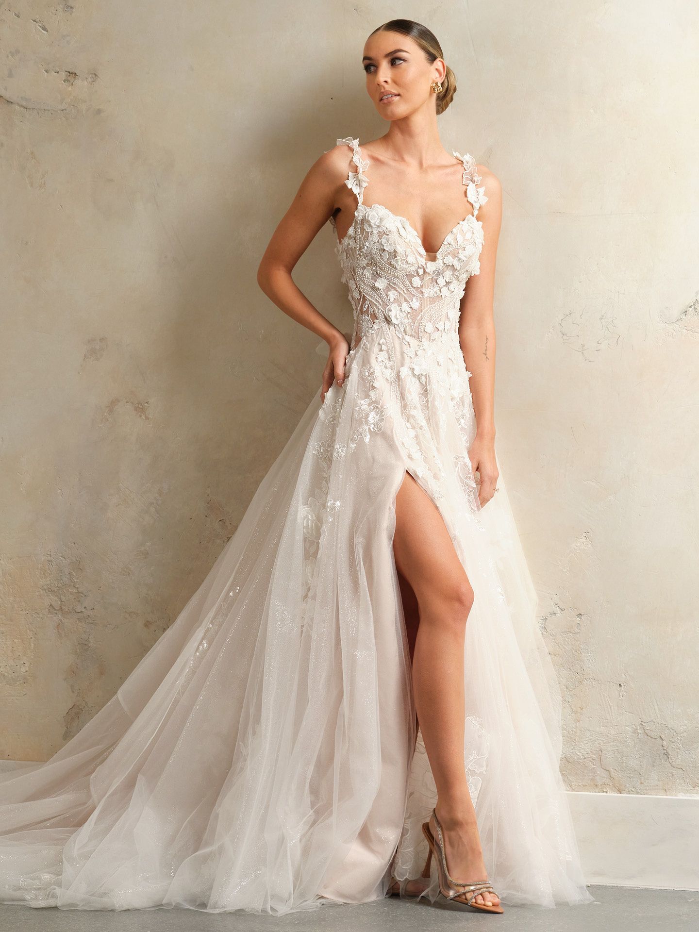 Woman in a white wedding gown with floral detail and a high slit, posing against a textured wall.