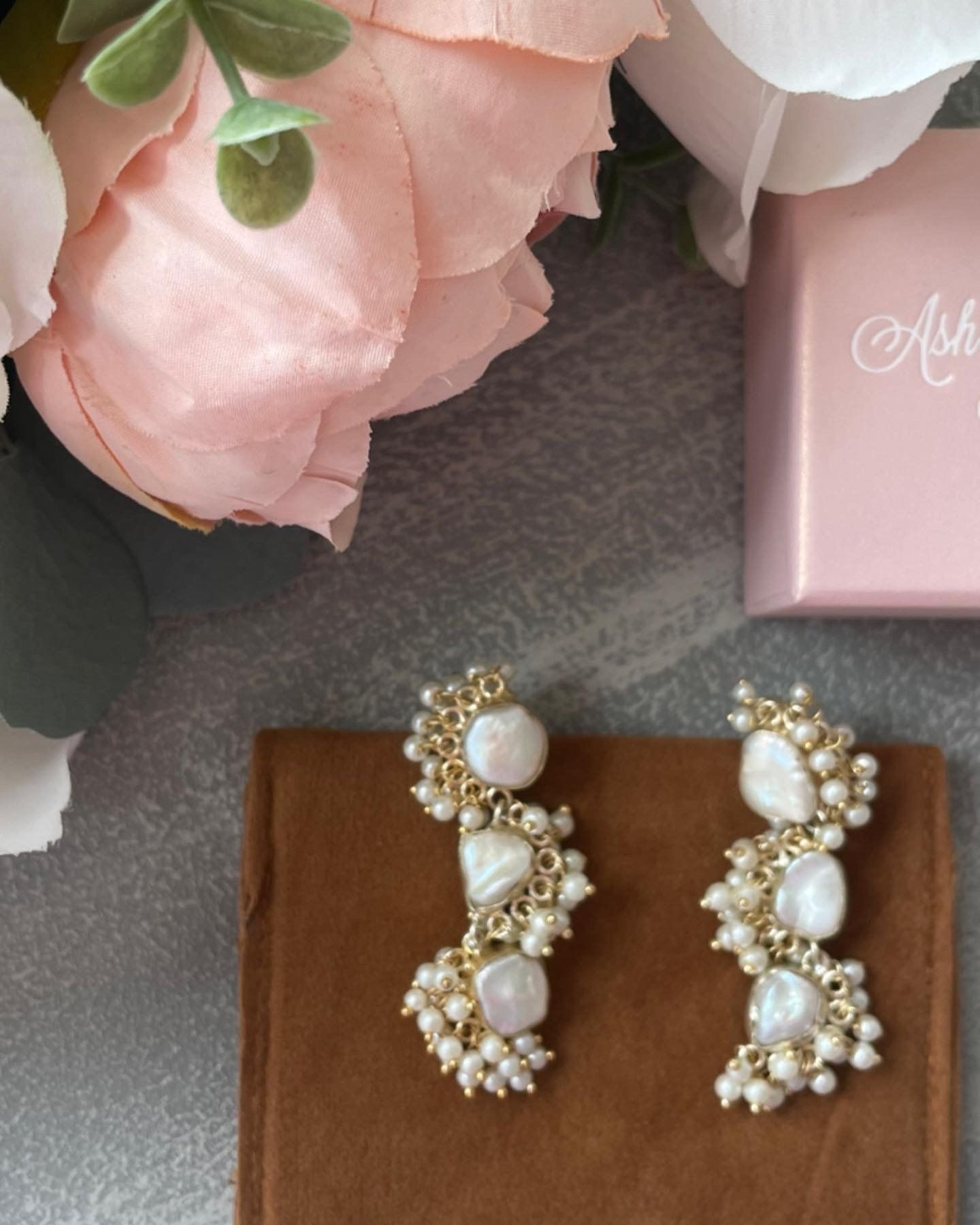 A pair of pearl earrings sit on a brown cloth next to a pink box
