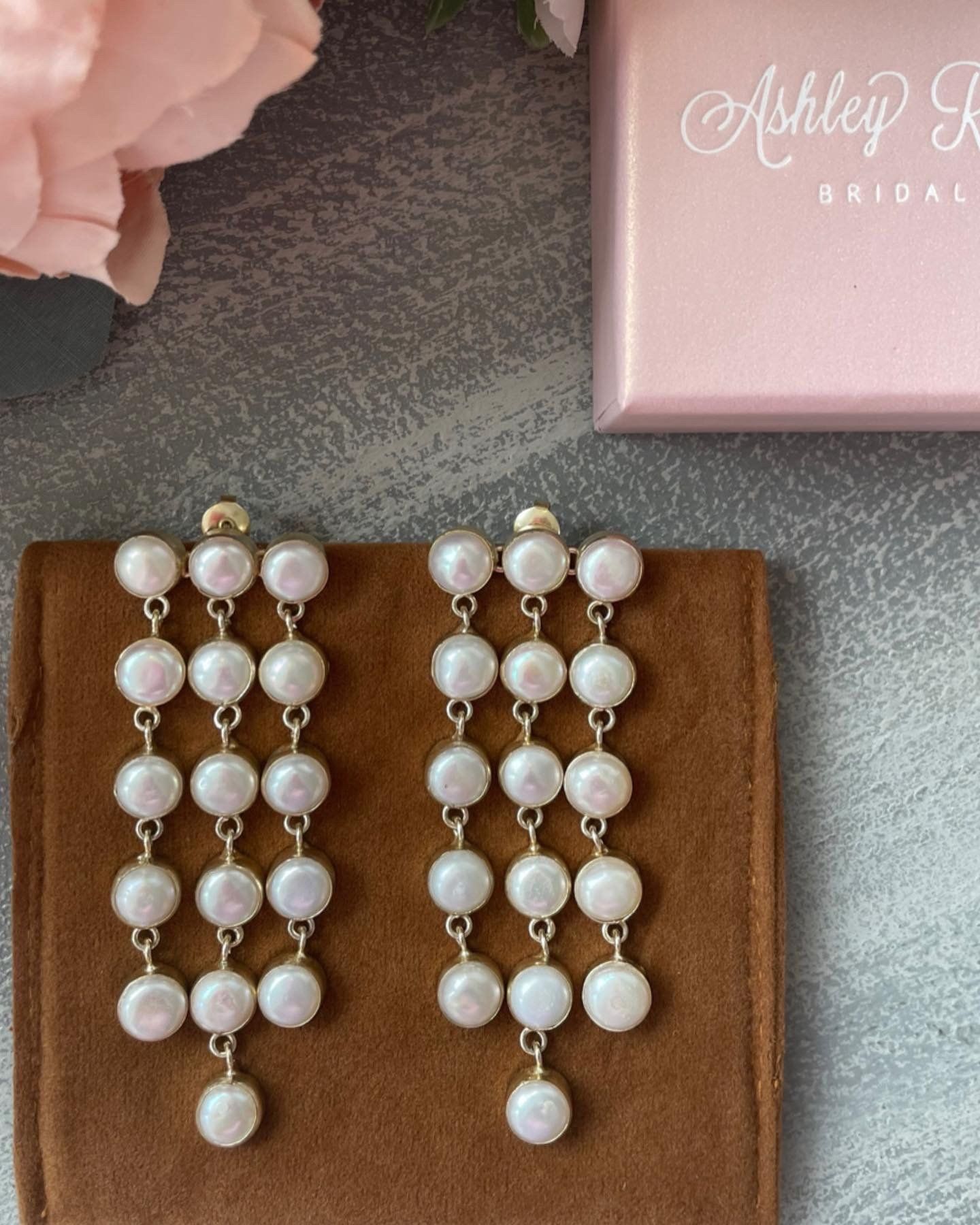 A pair of pearl earrings are sitting on a brown cloth next to a pink box