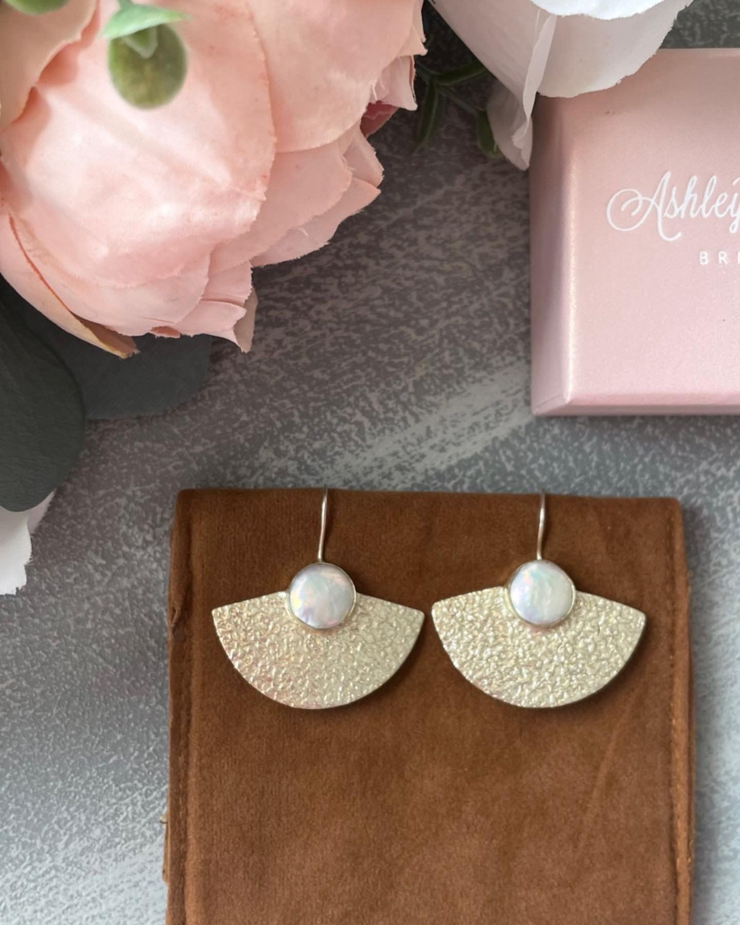 A pair of earrings are sitting on a brown cloth next to a pink box that says ashley