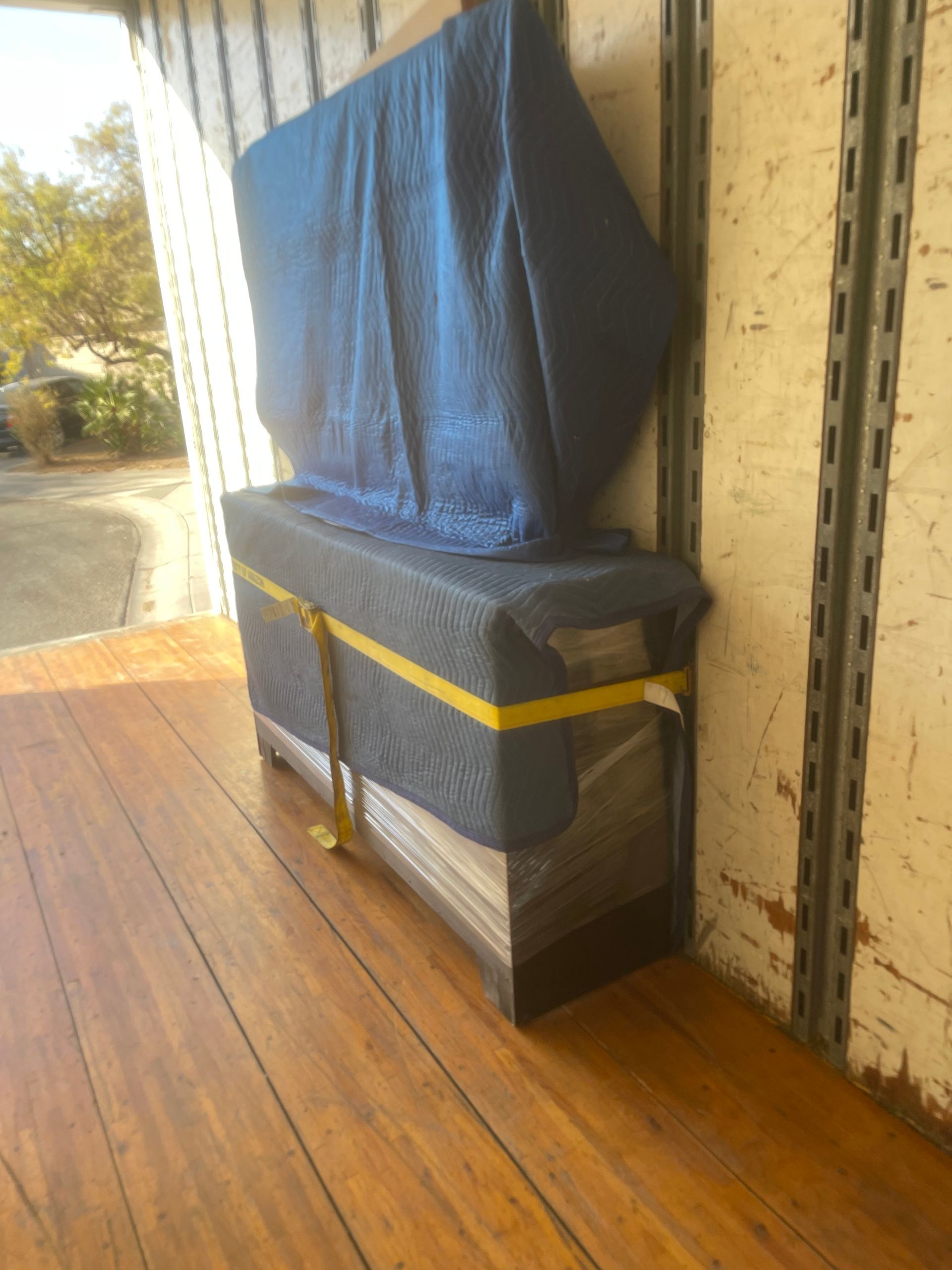 Television wrapped in blue moving blankets, secured with yellow straps inside a truck with wood flooring.