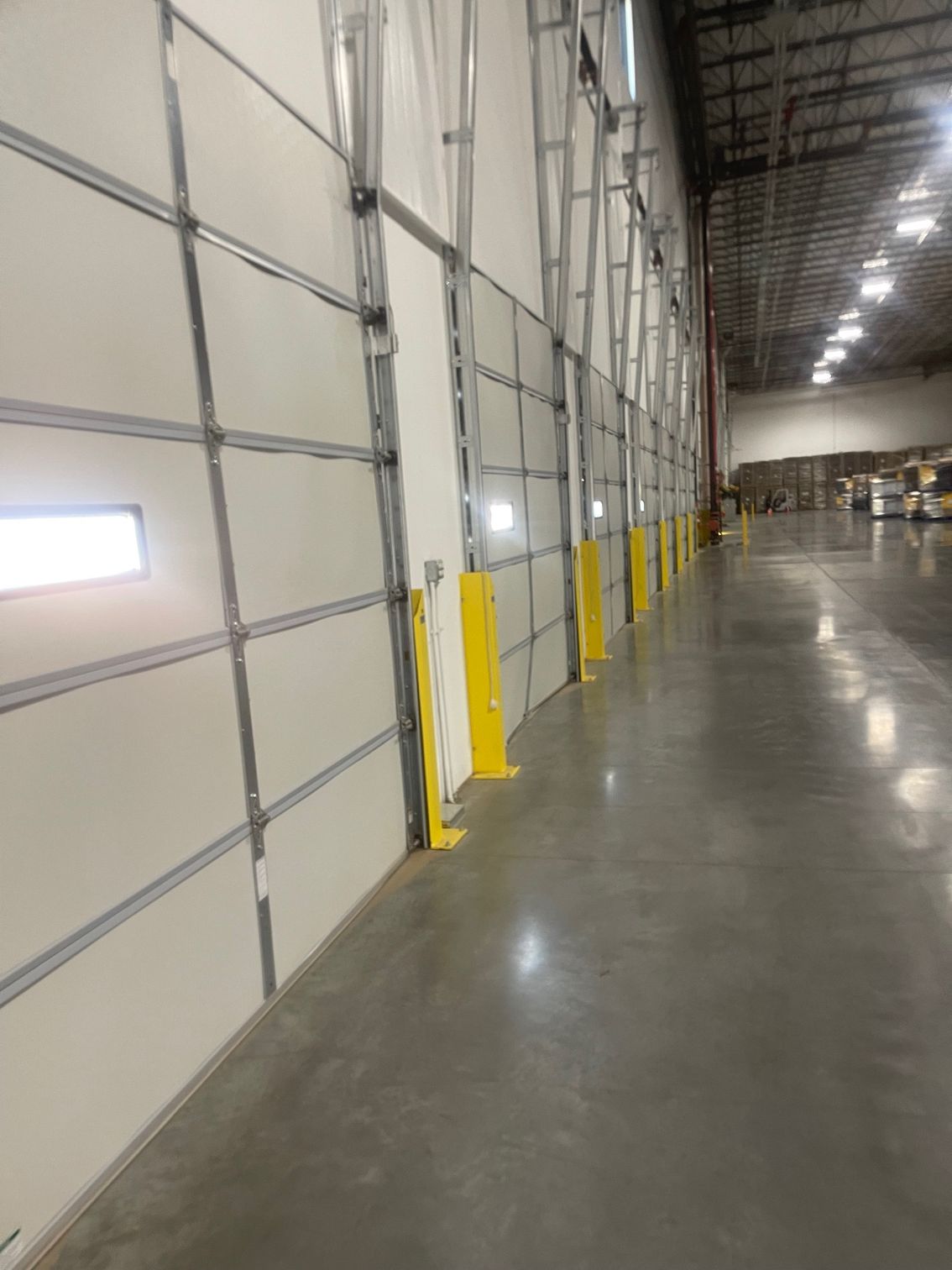 Inside a warehouse, a row of white, closed overhead doors along a wall with yellow safety posts.
