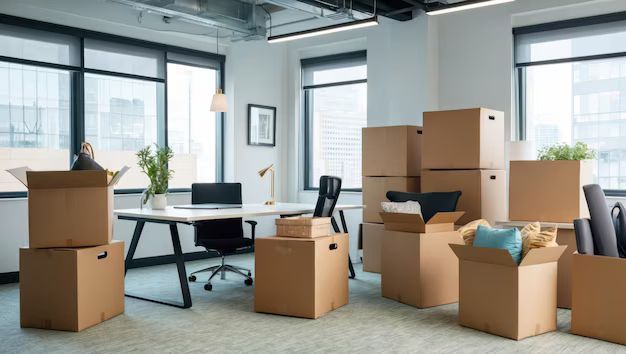 Office space filled with cardboard moving boxes. Desk with chair, window views.