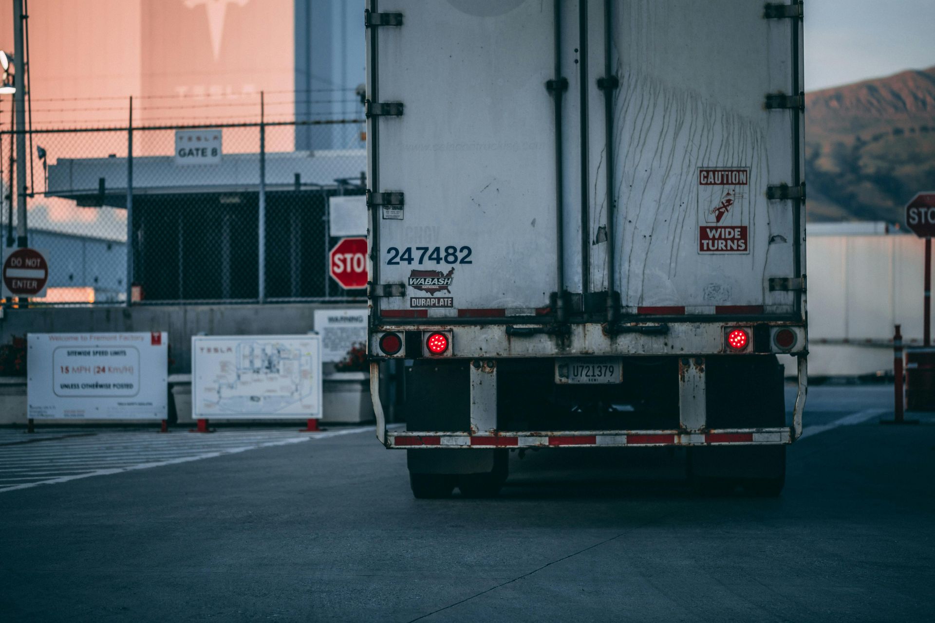 Rear view of a semi-truck approaching a gate. Red taillights, stop sign, Tesla sign visible in the background.