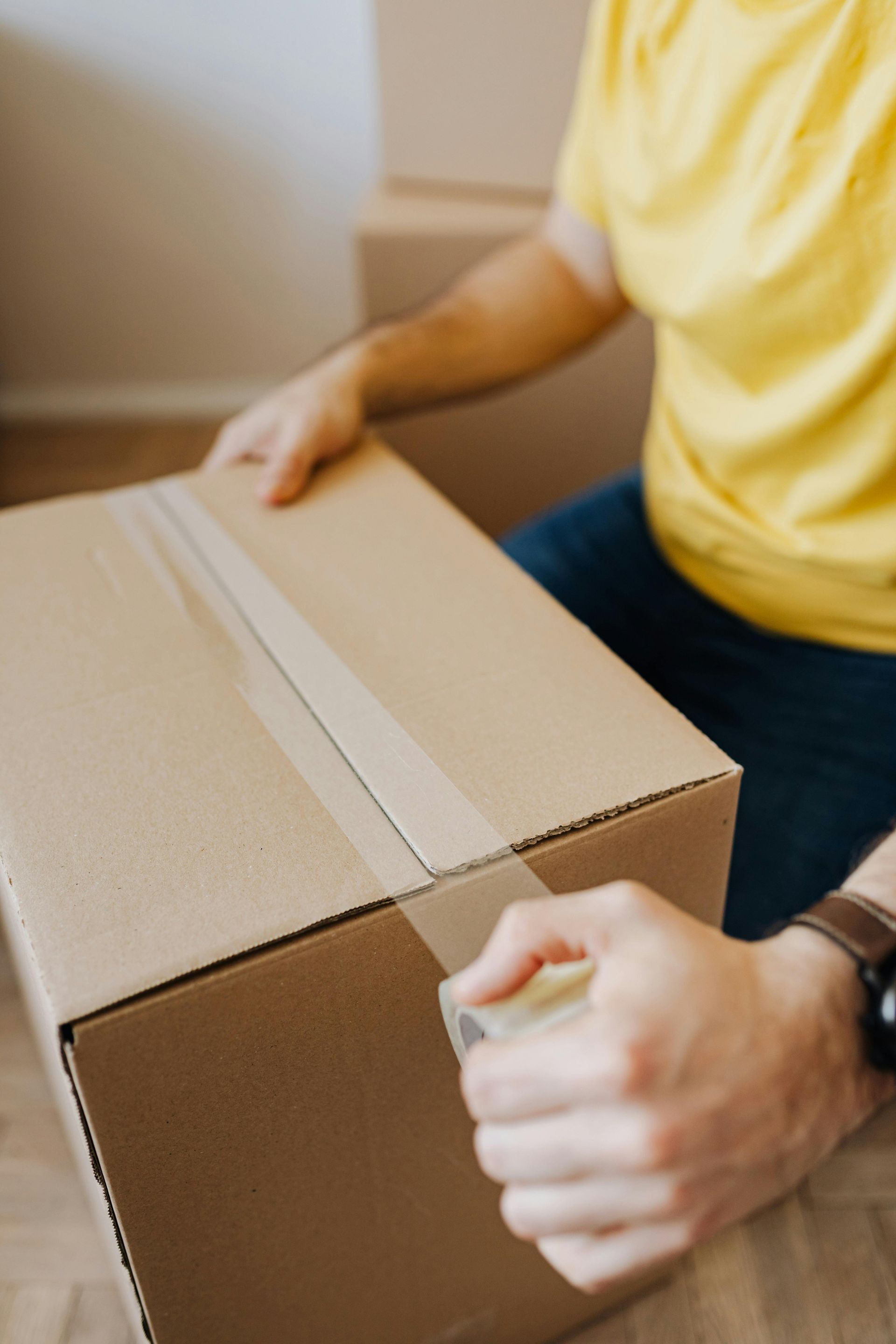 Person sealing a cardboard box with clear tape; in a room, wearing yellow shirt, jeans, and a watch.