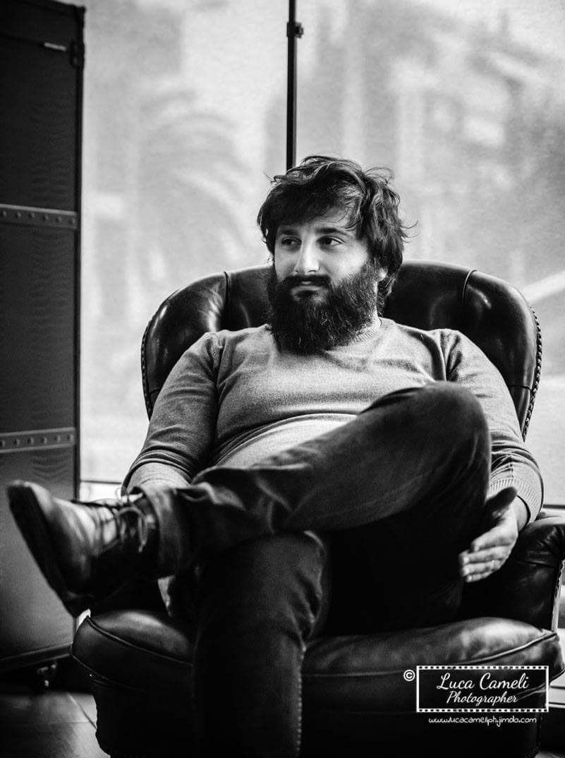 Uomo con barba folta, seduto su una poltrona di pelle, in posa rilassata, gambe incrociate.