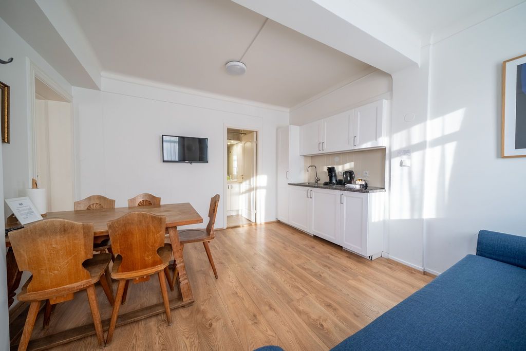 A studio apartment with a wooden dining table and chairs, a kitchenette with white cabinets, and a blue sofa.