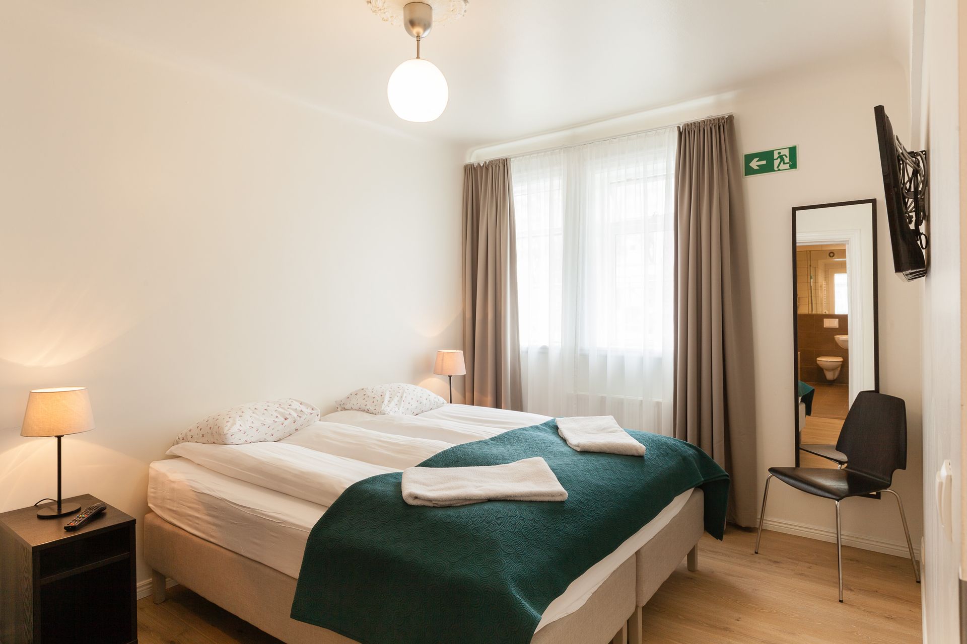 A hotel room with a queen bed featuring a green quilt, a nightstand, a mirror, a chair, and a bright window with curtains.