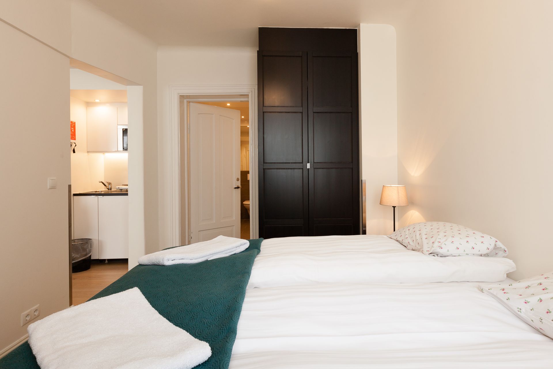 A bedroom with a bed in the foreground, a dark cabinet behind it, and a view into a small kitchen and a white doorway.