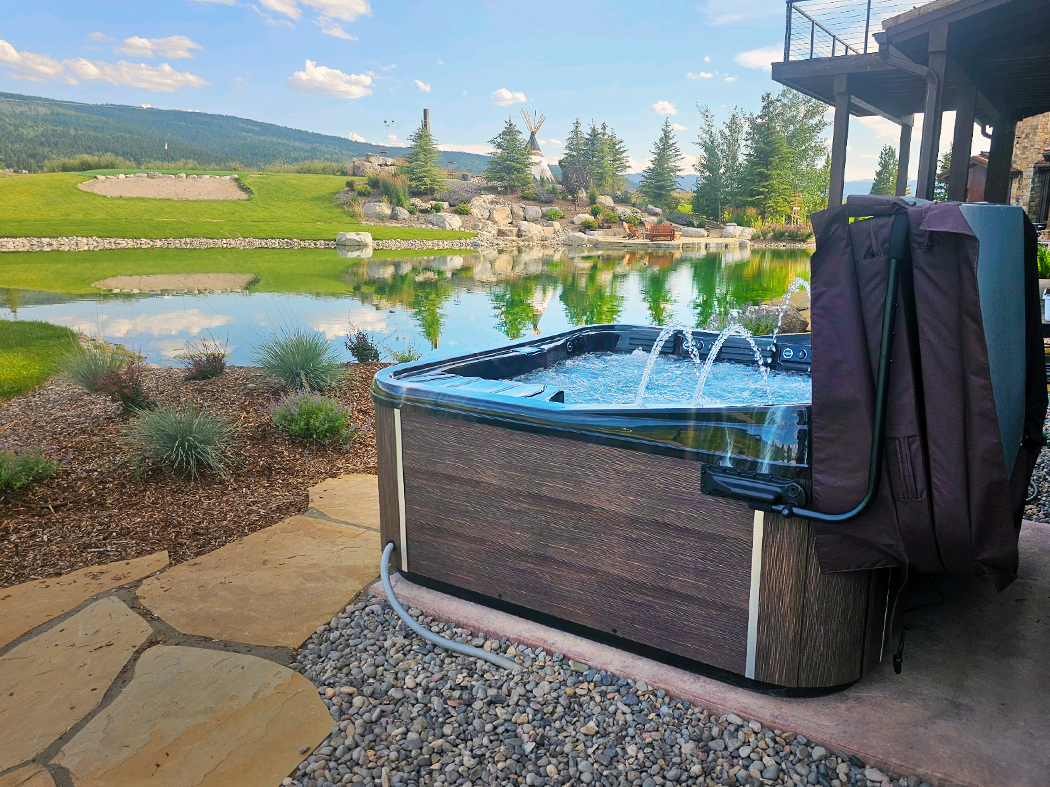 Hot tub with water fountain in a scenic outdoor setting, brown siding, stone patio, lake, and hills in background.