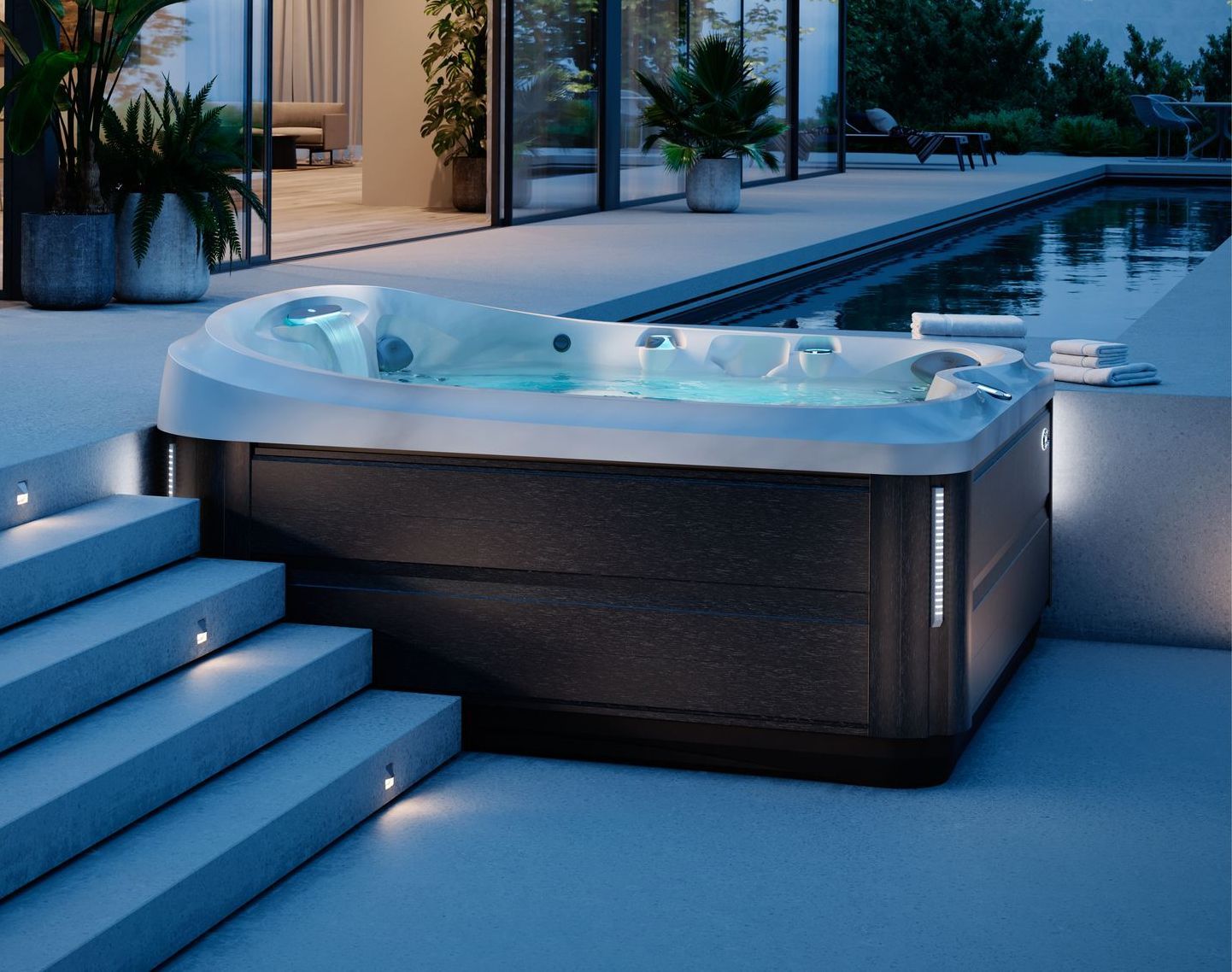 Hot tub on a patio with steps, lit up at dusk. Blue water, dark exterior, pool in the background.