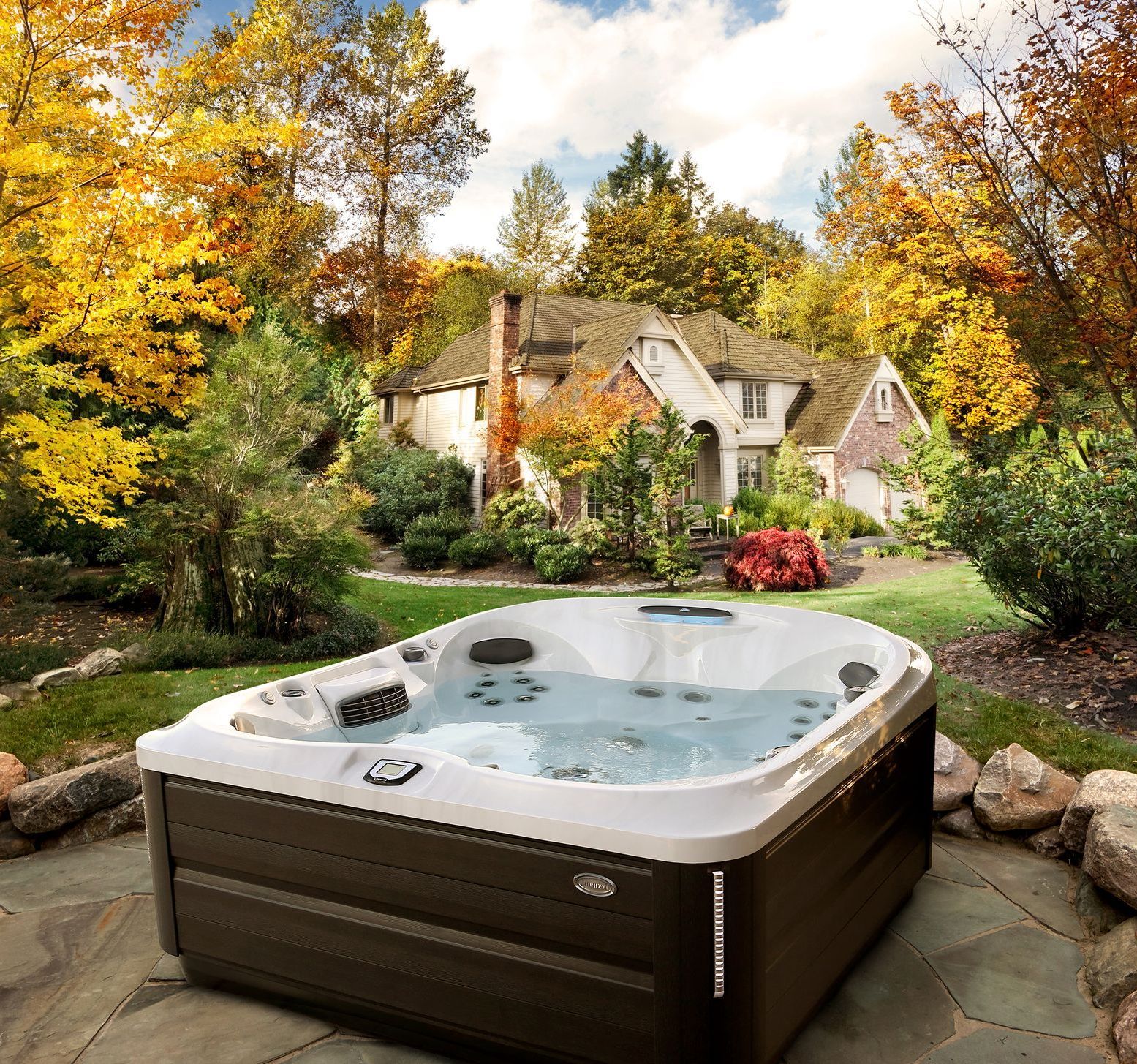 Hot tub on patio in front of a house with fall foliage; brown and white exterior, green lawn.
