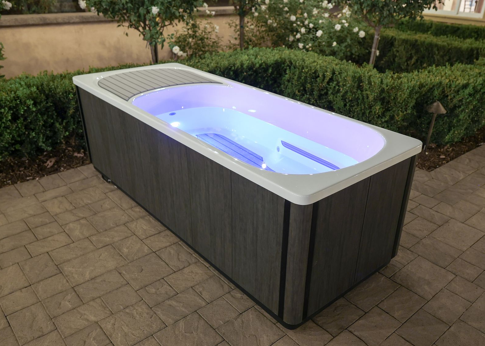 Rectangular hot tub with gray siding, lit with blue lights, on a brick patio.