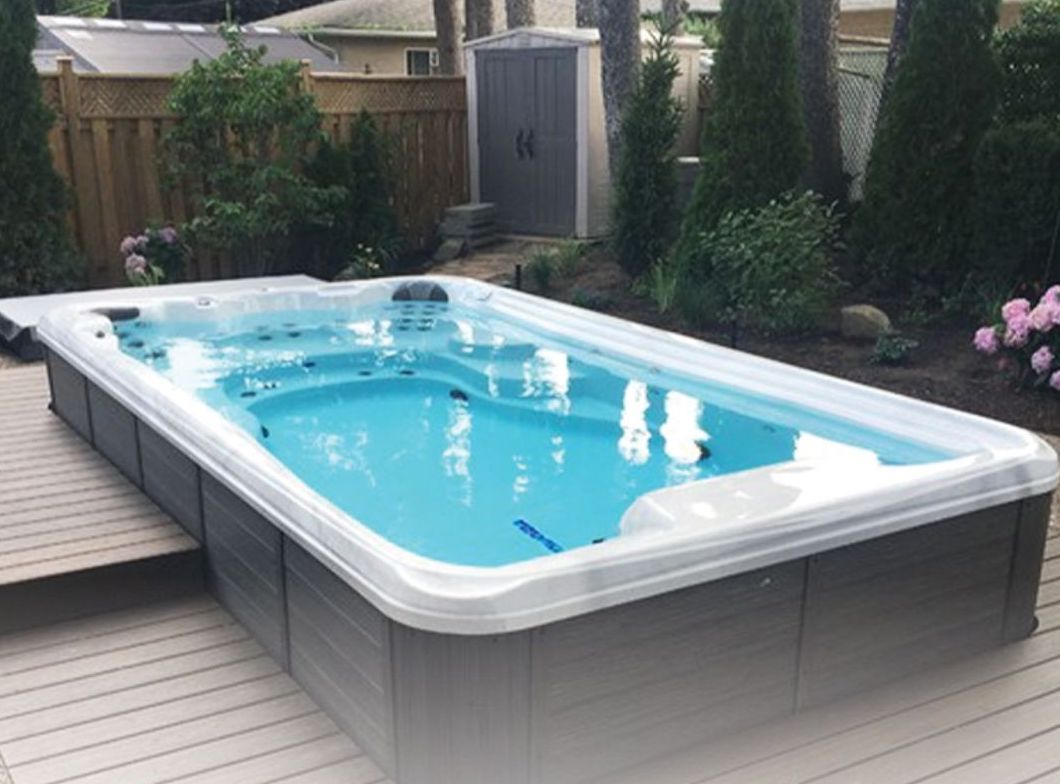 Swim spa with gray surround, blue water, on a wooden deck, surrounded by greenery and a fence.