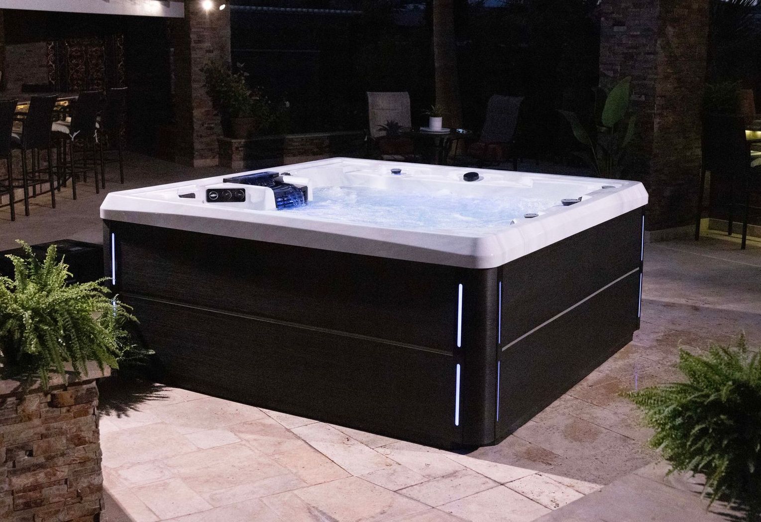 Square hot tub lit with blue lights on a patio at night.