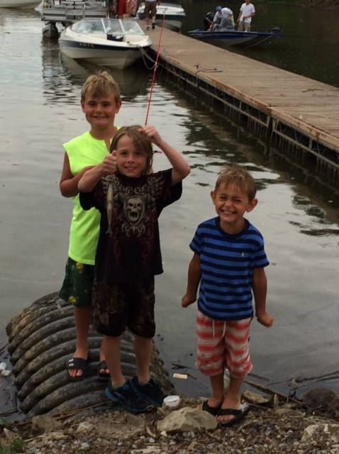 Three young boys are fishing near a dock with a boat in the background