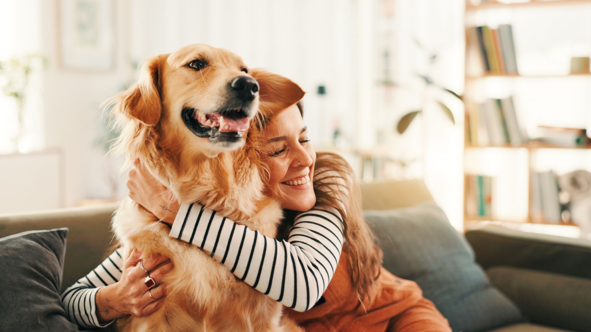 A woman in a striped long-sleeve shirt hugs a happy golden retriever while sitting on a couch.