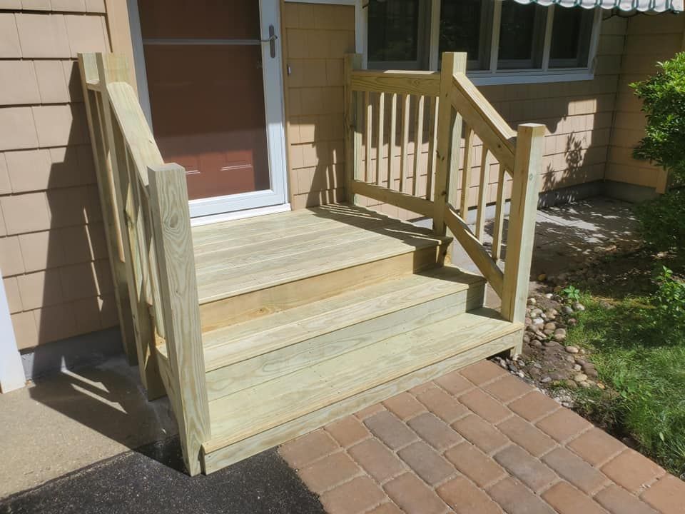 New wooden front steps with railings leading to a house entrance, adjacent to a paver walkway.