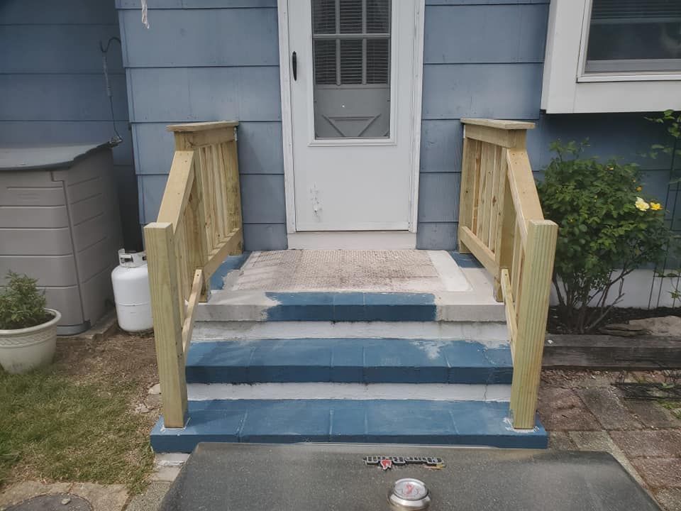 A set of three outdoor stone steps with new wooden railings leading up to a white door on a blue-sided house.