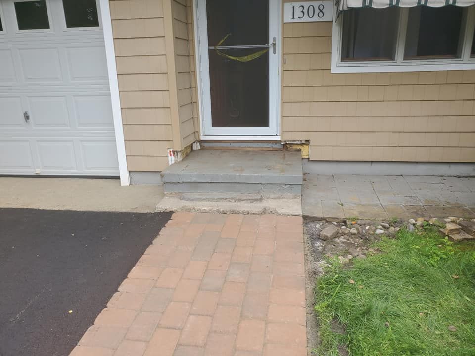 A front entrance featuring a brick paver walkway leading to two concrete steps and a tan house with a white front door.