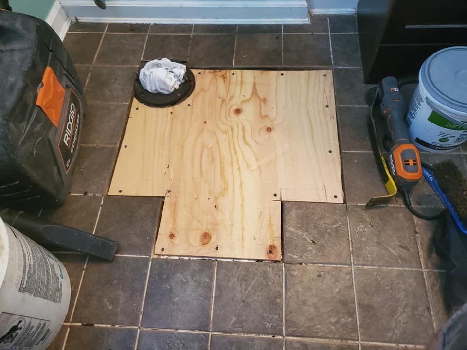 A floor patch with a piece of plywood installed within a tiled floor, surrounded by construction tools and materials.