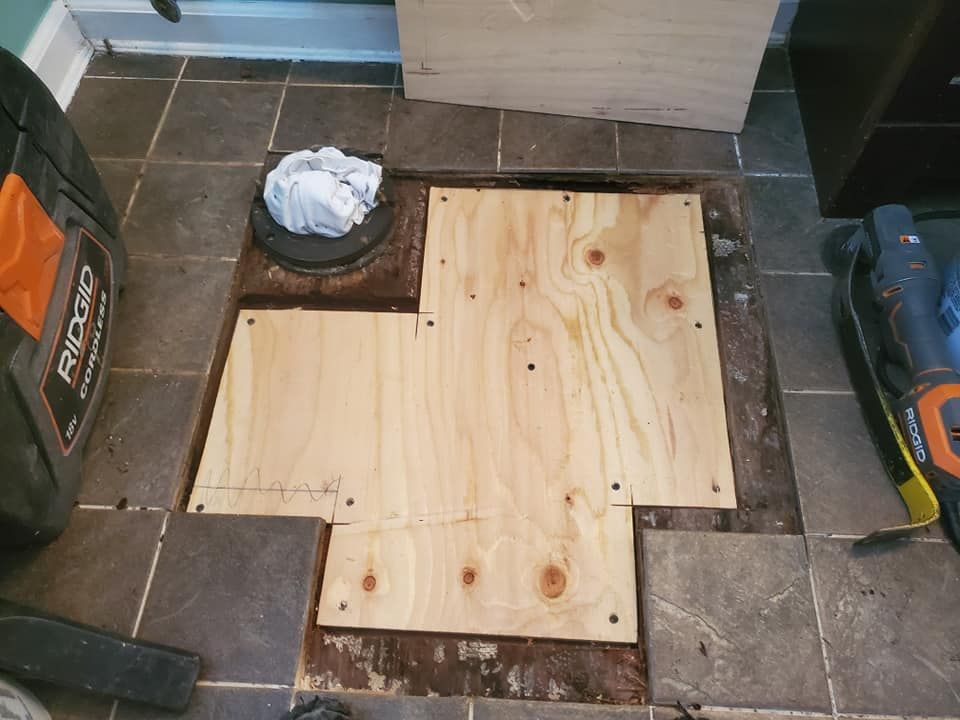 A hole cut into a tiled floor exposing subflooring, with new plywood patches installed around a toilet drain pipe.
