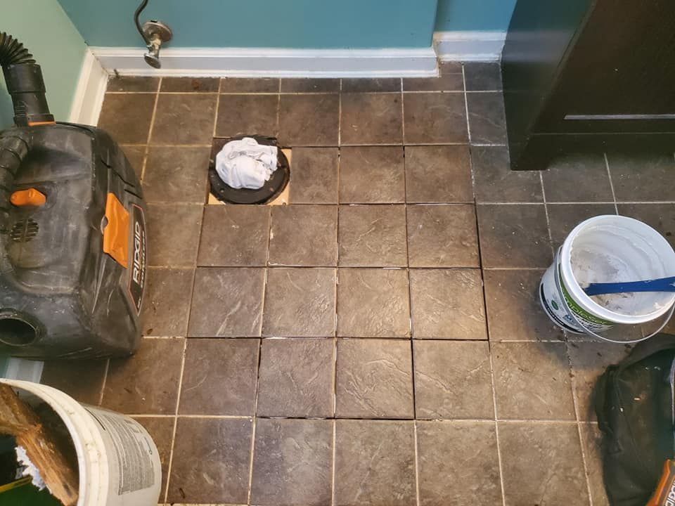 A bathroom floor with brown square tiles, a toilet flange with a rag in the drain, a vacuum, and a bucket of grout mix.