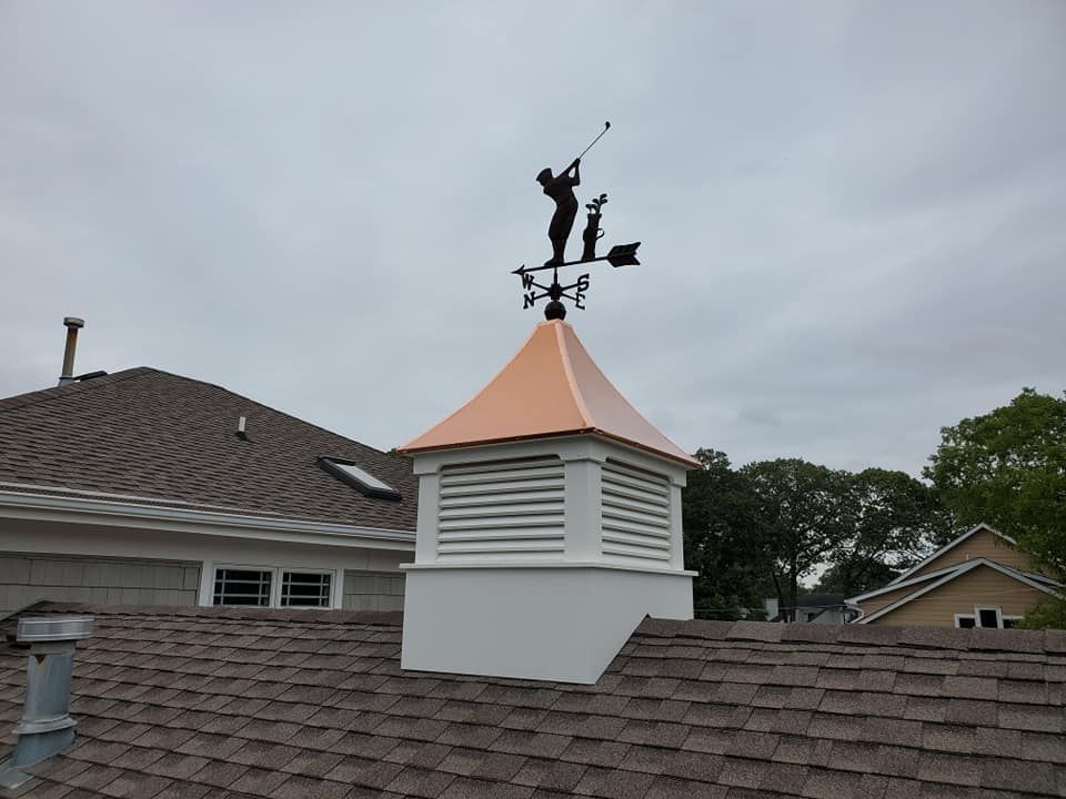 A white square cupola with a copper roof and a golfer weathervane, mounted on a brown shingled residential roof.