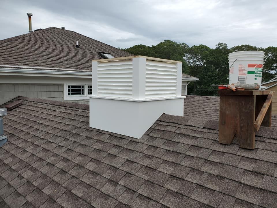 A white custom chimney shroud sits on a brown shingled roof next to a wooden sawhorse and a bucket.