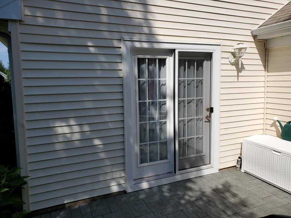 A light-colored house exterior featuring a glass sliding door with a white frame and a screen, set on a stone patio.