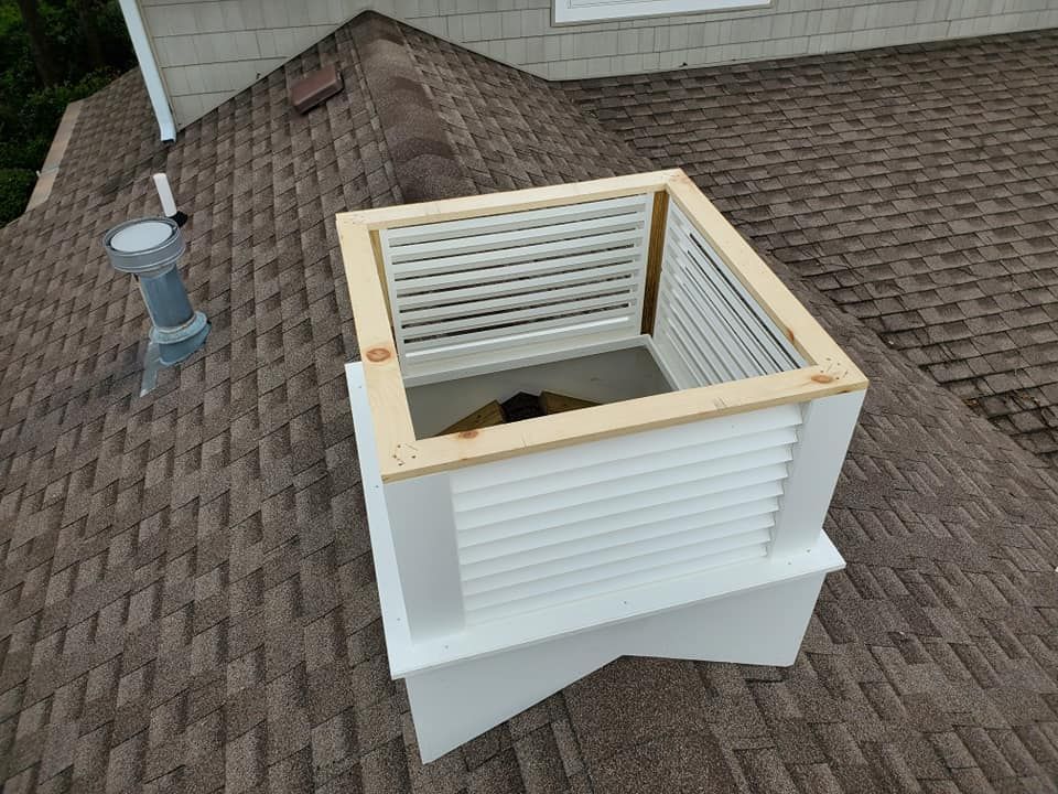 A square, white louvered cupola with a wooden top frame sits unfinished on a brown shingled roof.