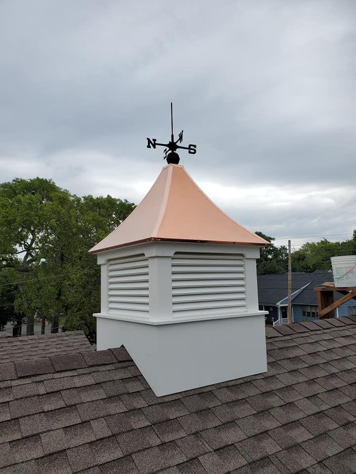 A square white wooden cupola with a copper roof and a black weathervane sits on a shingled roof under a cloudy sky.